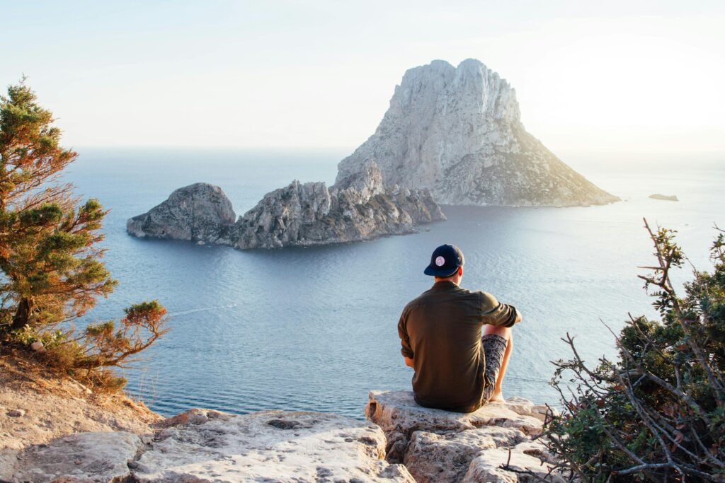 Man enjoys a scenic view of Es Vedrà at sunset from a cliff in San Juan Bautista, providing a perfect summer escape and he is playtech's vip member